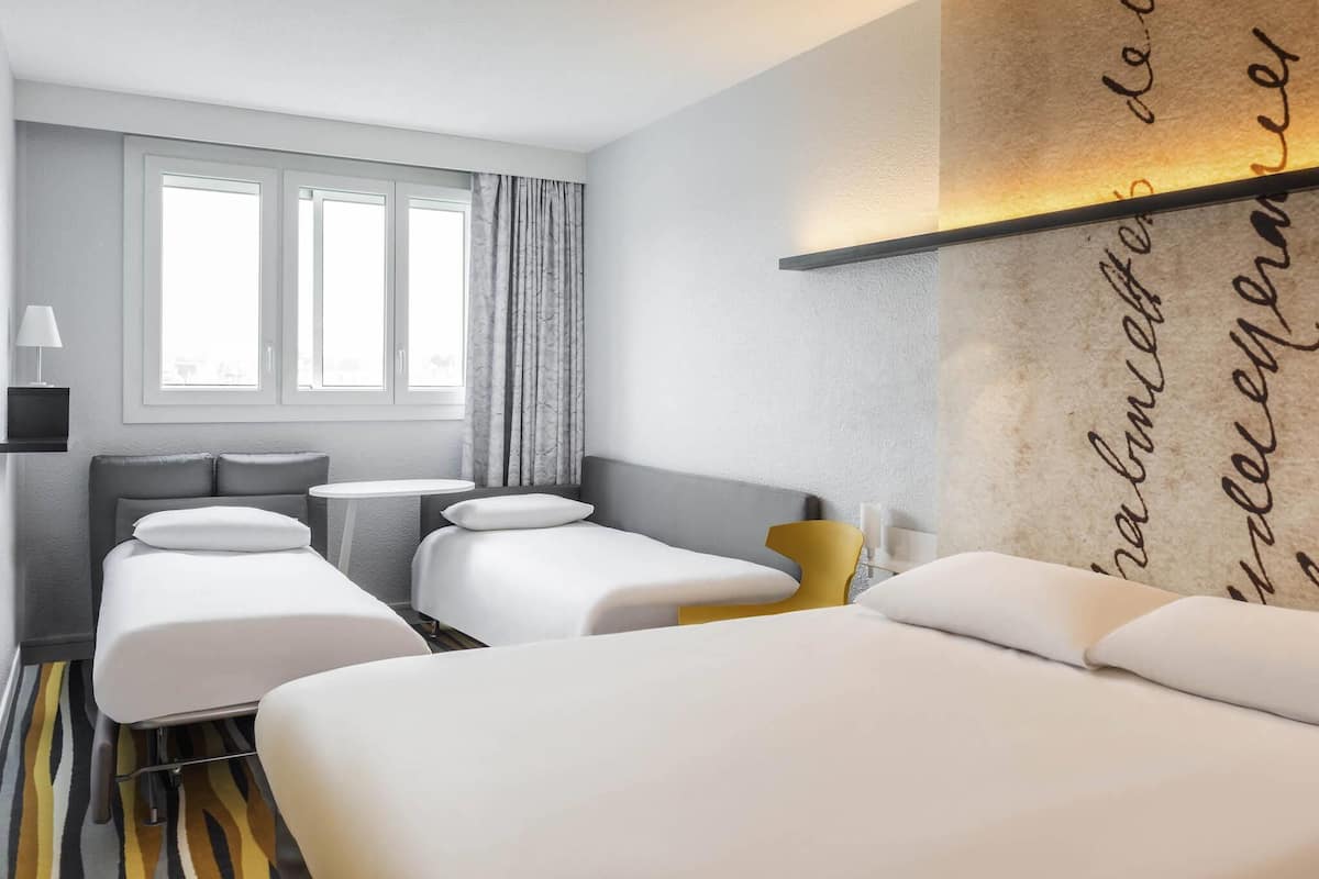 Habitación de hotel con dos camas, una mesita, una lámpara y una ventana con cortinas.
