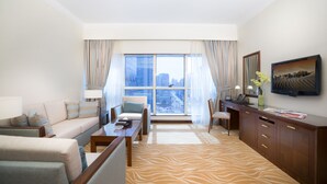 A hotel room with a sofa, armchairs, a desk, a television, and a large window with a city view. - Majlis Grand Mercure Residence Abu Dhabi (Abu Dhabi)