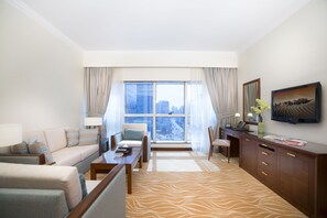 Habitación de hotel con sofá, sillones, escritorio, televisor y un amplio ventanal con vistas a la ciudad.
