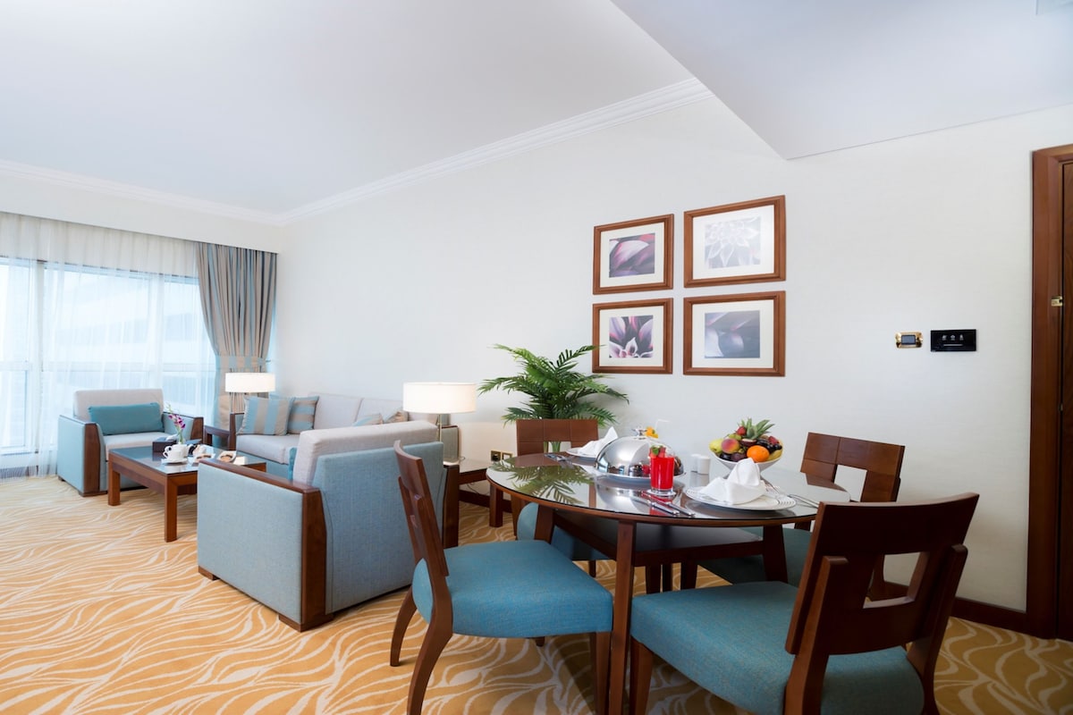 Habitación de hotel con sala de estar, una mesa de comedor y sillas. Hay cuatro cuadros en la pared, una planta en maceta y una ventana con cortinas.