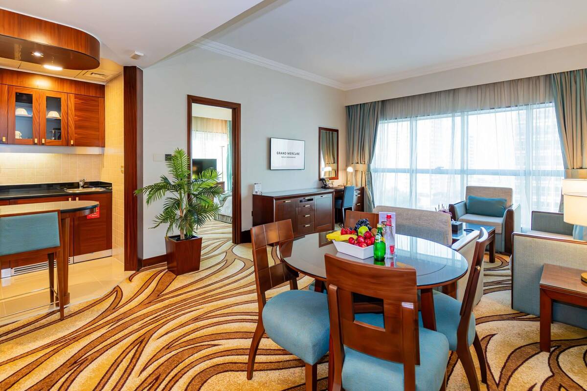 Habitación de hotel con comedor, cocina, sala de estar con sofá y sillones, y un amplio ventanal con cortinas.