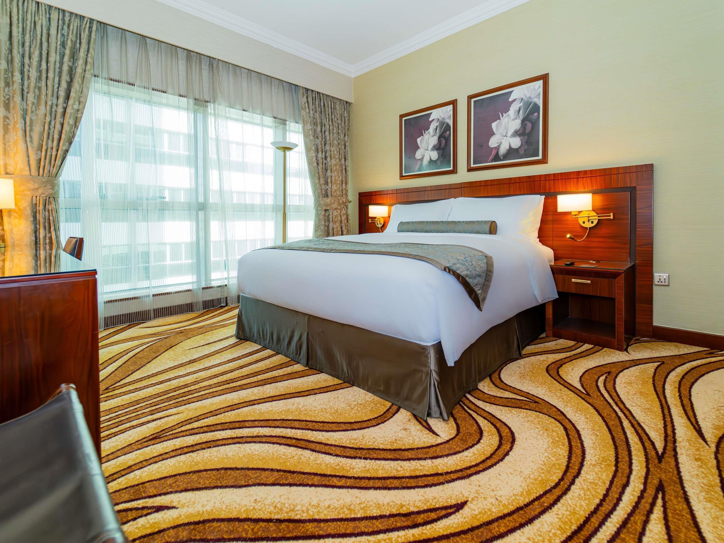 A hotel room with a large bed, a wooden headboard, two wall-mounted lamps, and a large window with sheer curtains.