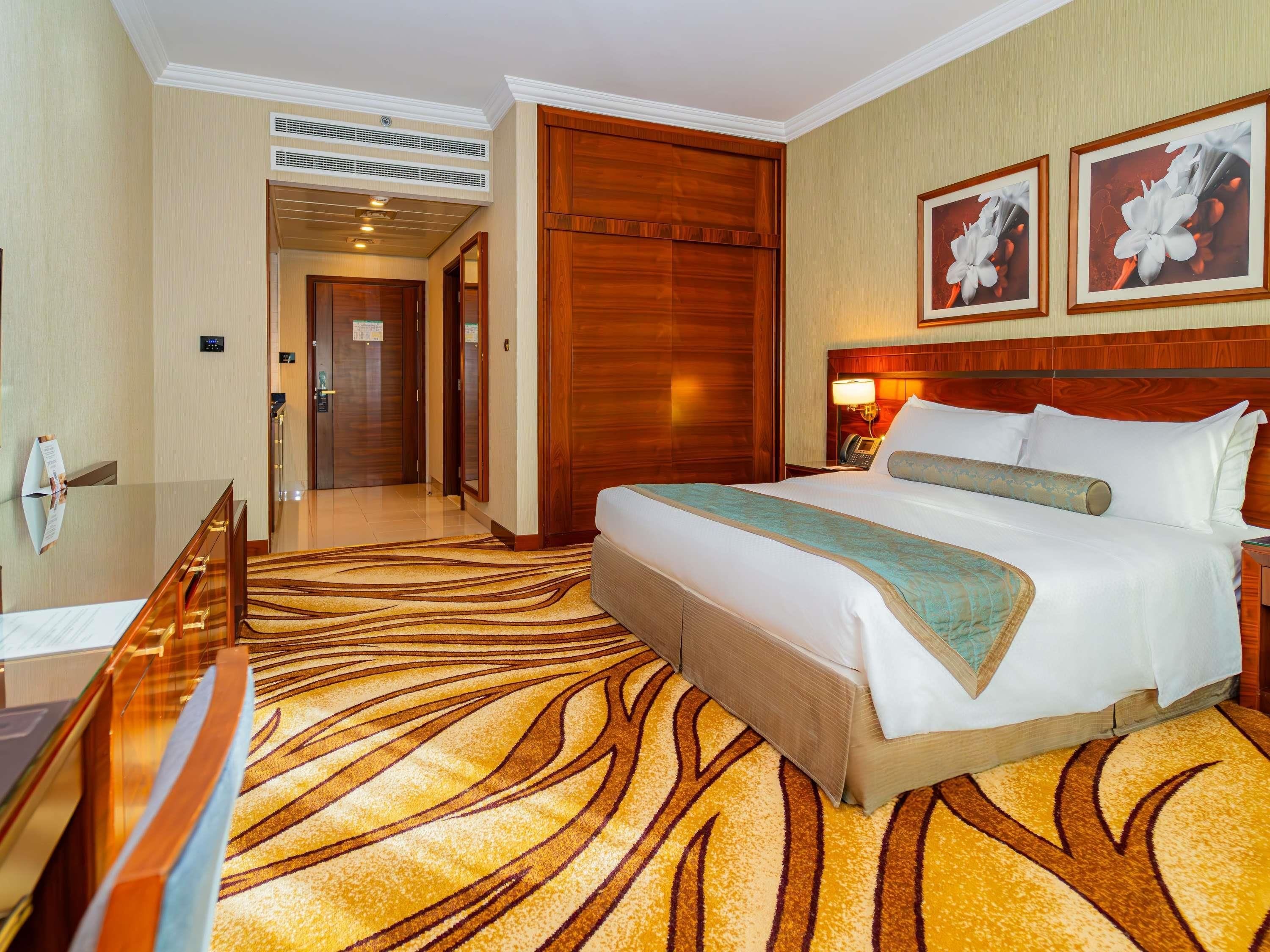 A hotel room with a bed, a wooden headboard, a nightstand with a lamp, a desk with a chair, and a carpet with a leaf pattern.