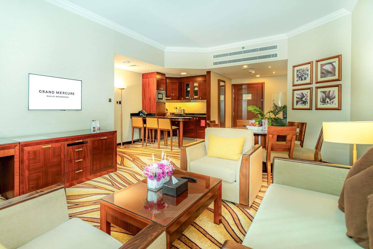 Habitación de hotel con sala de estar, cocina y comedor. Hay un televisor de pantalla plana en la pared, una mesa de centro con un jarrón de flores y un sofá.