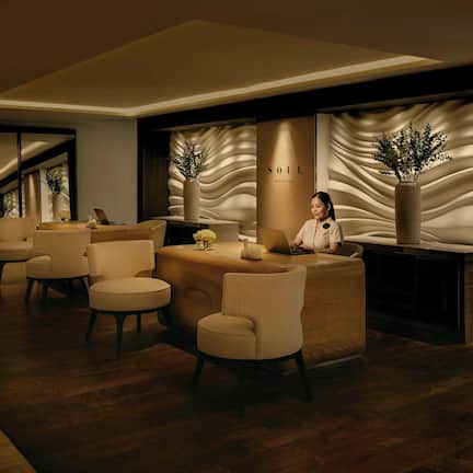 A modern reception area with a receptionist, a reception desk, several chairs, and decorative plants.