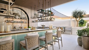 A bar area with a tiled backsplash, hanging shelves with bottles, and wicker bar stools. - Sheraton Grand Hotel, Dubai (Dubai)