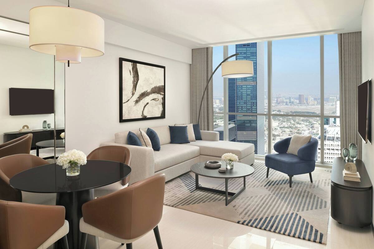 A modern living room with a sofa, armchairs, a coffee table, and a dining table. There is a large window with a city view.