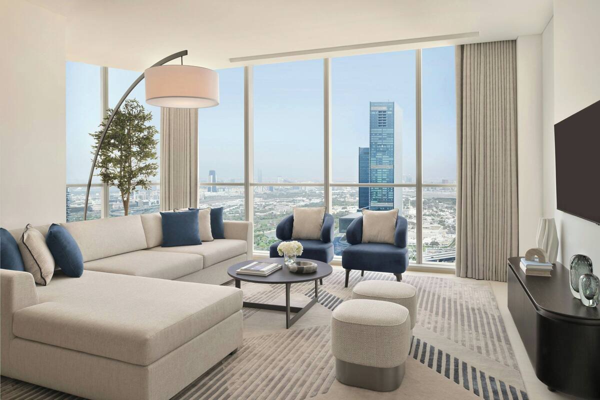 A modern living room with a sofa, armchairs, a coffee table, and a TV. A large window offers a city view.