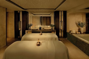 A modern spa room with two large beds, a massage table, a large bathtub, and a mirror.