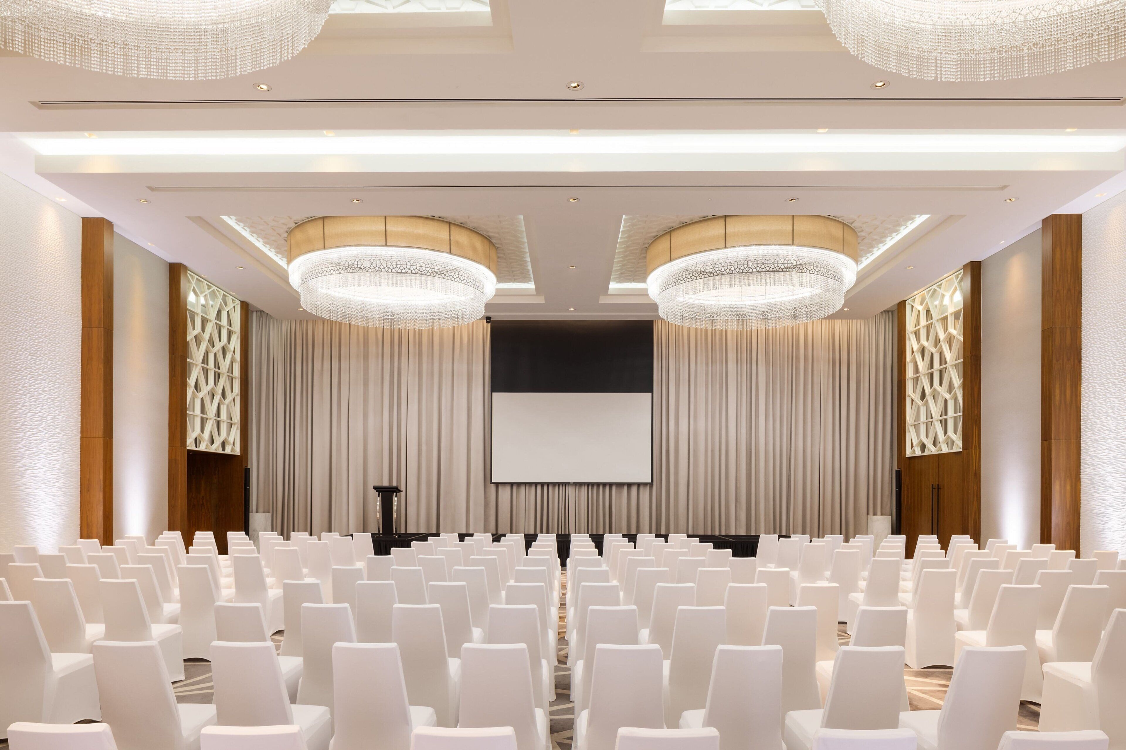 A conference room with rows of white chairs, a large screen, and two chandeliers.