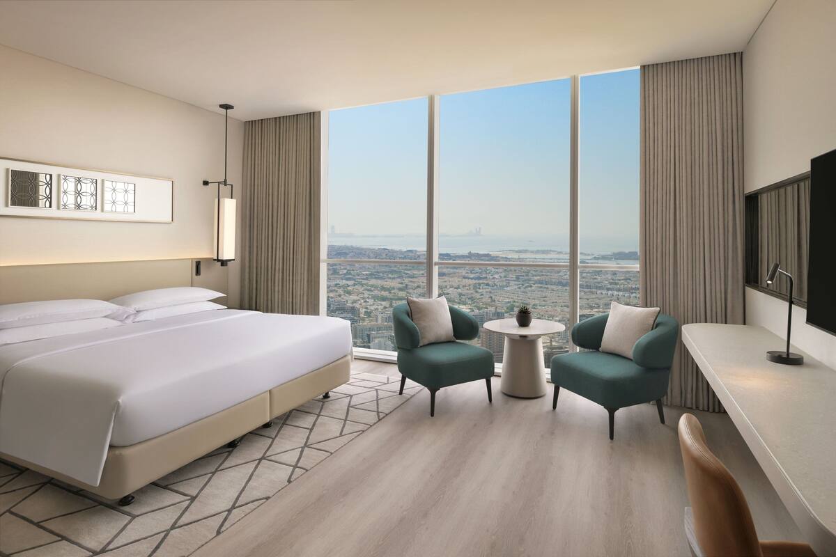 A modern hotel room with a large bed, two armchairs, a small table, and a TV. The room has large windows offering a city view.