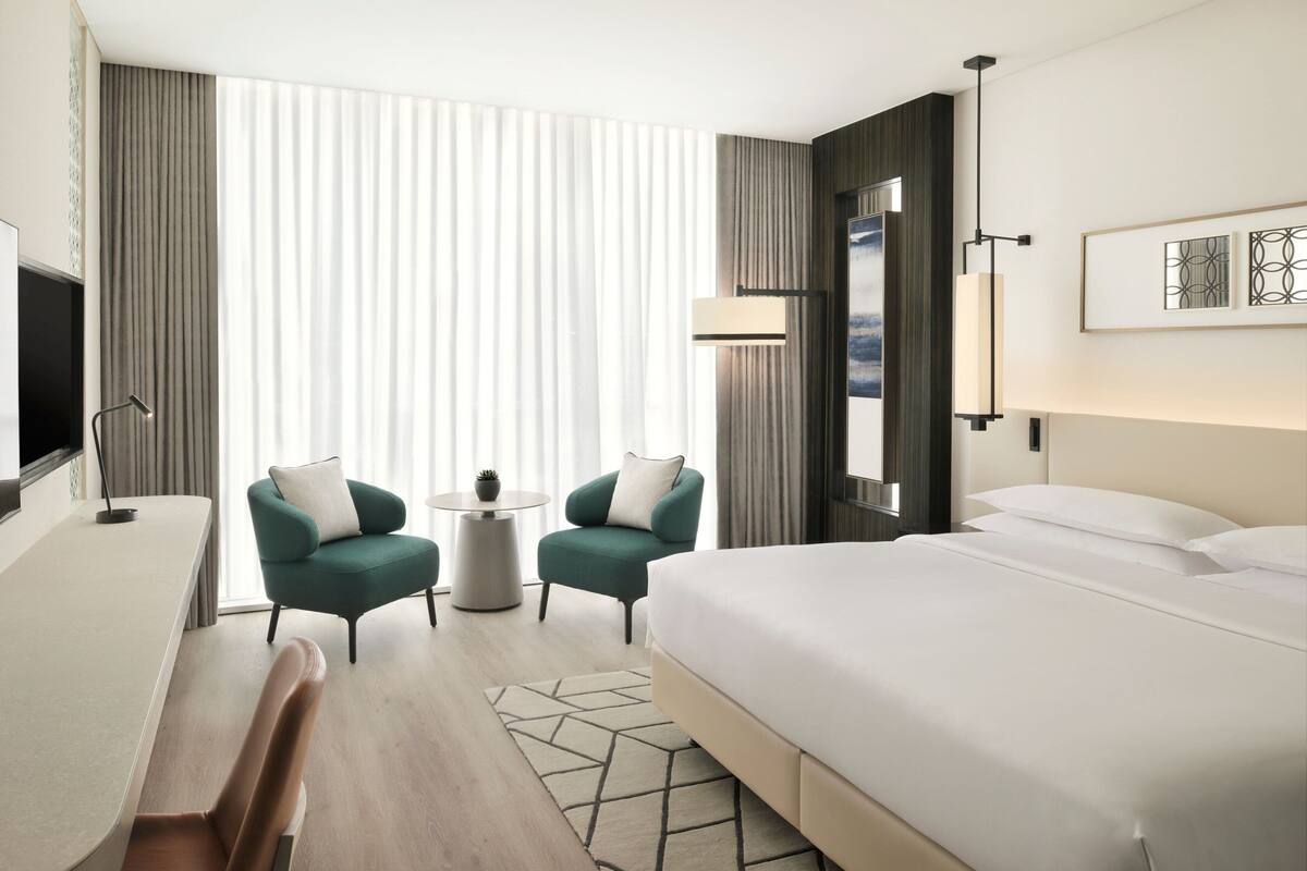 A modern hotel room with a large bed, a desk, two armchairs, a small table, and a TV.