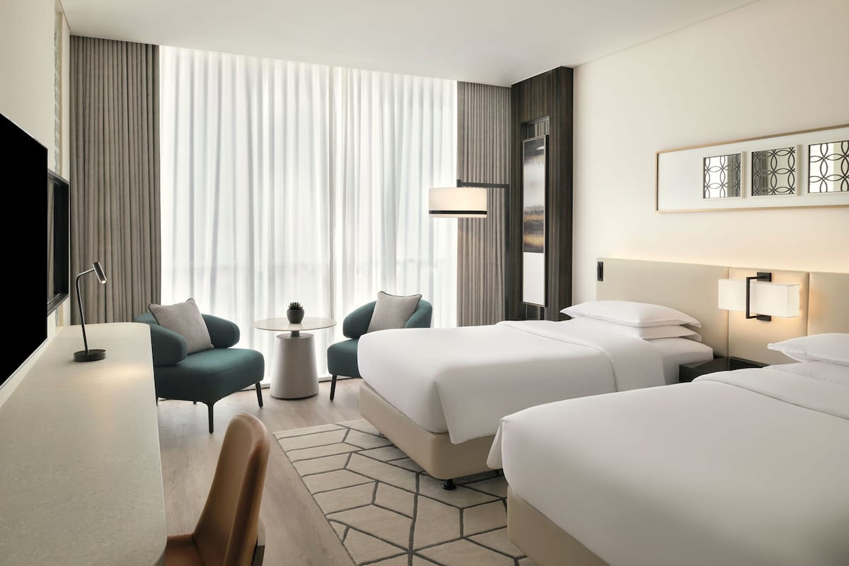 A hotel room with two beds, a desk, a chair, a lamp, and a large window with curtains.