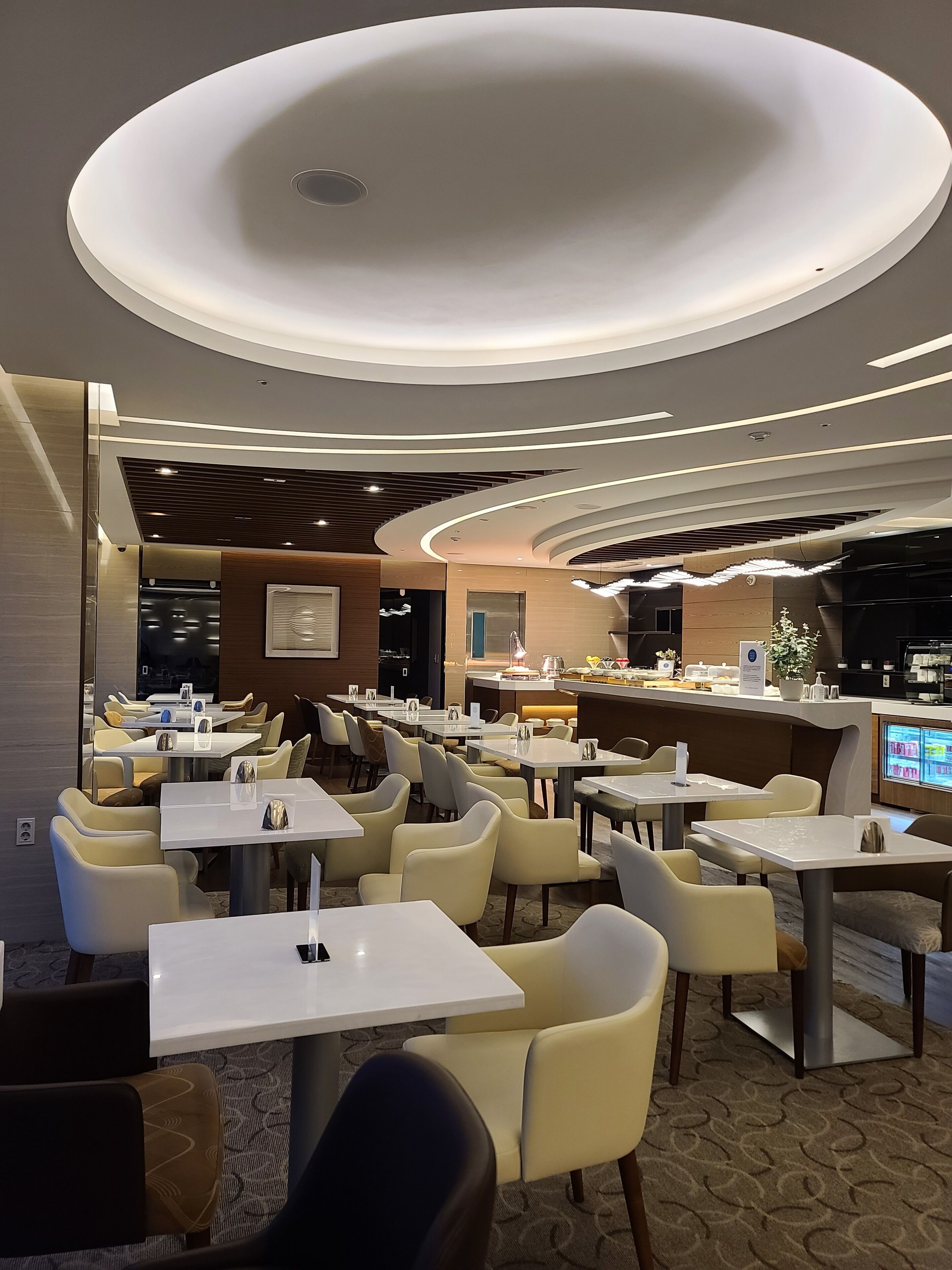 A modern restaurant with round tables and chairs, a counter with a digital menu display, and a ceiling with circular lighting.