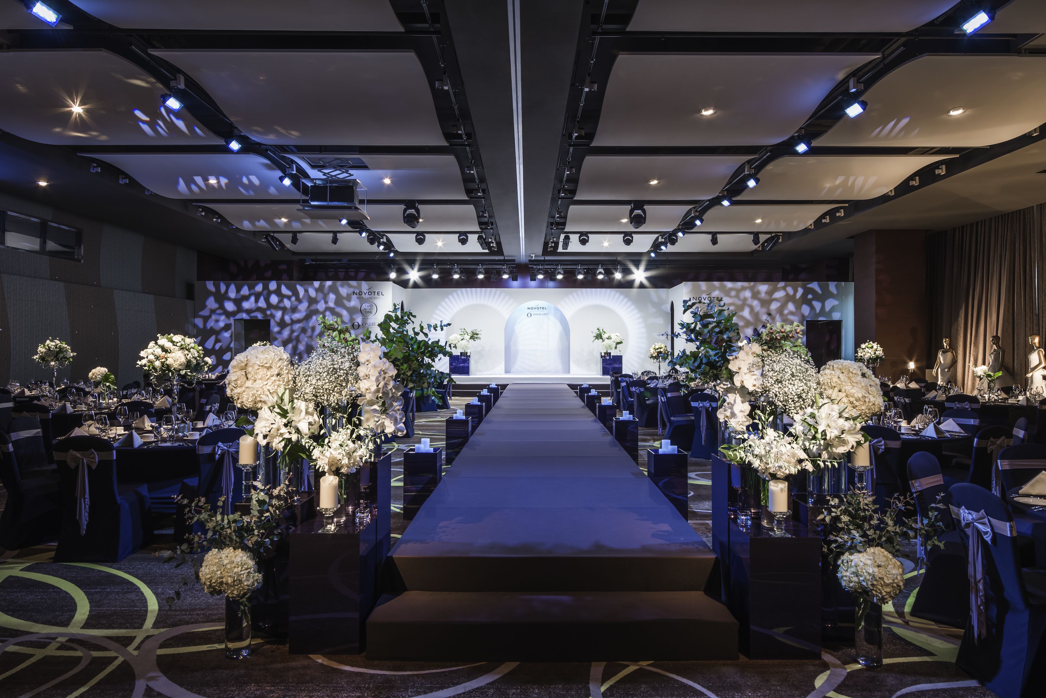 A hotel ballroom with a long, dark carpeted aisle leading to a stage, flanked by tables with floral centerpieces.