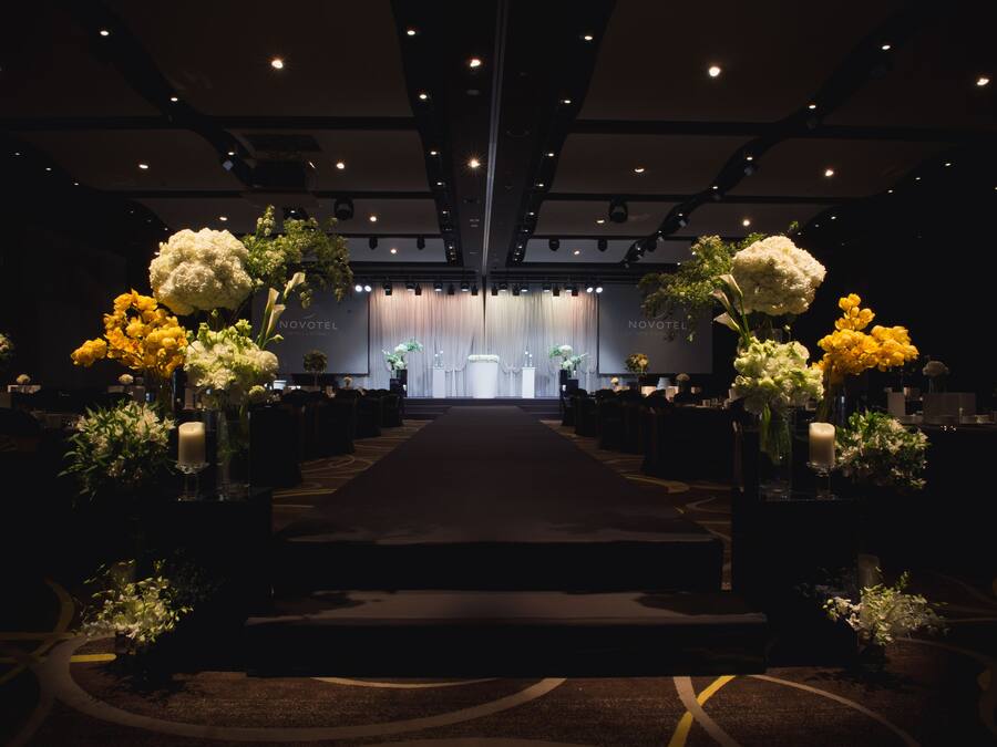 A hotel interior with a central aisle leading to a stage, flanked by floral arrangements and seating.