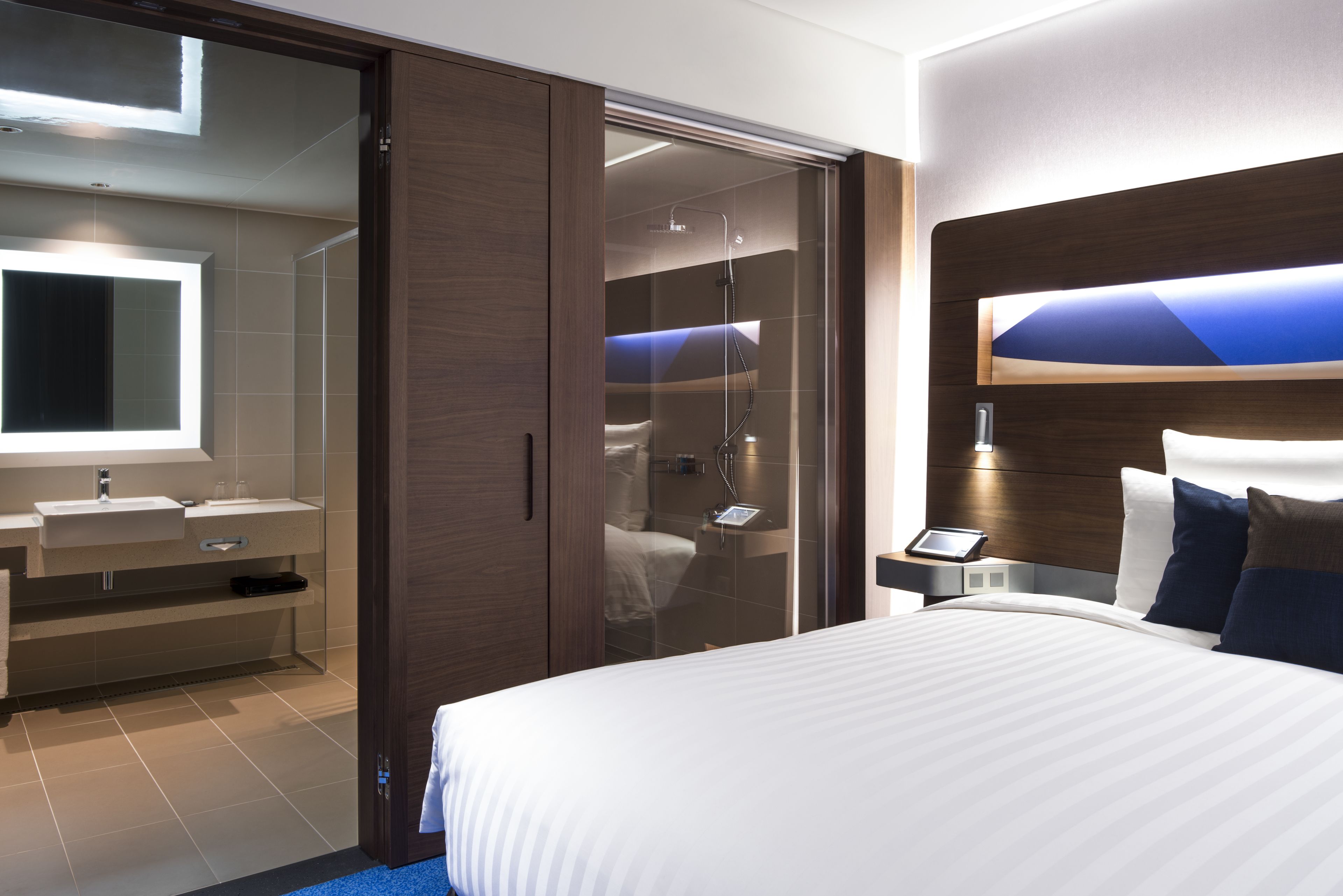 A modern hotel room with a large bed, a bathroom with a glass shower, a vanity with a mirror, and a bedside table with a lamp.