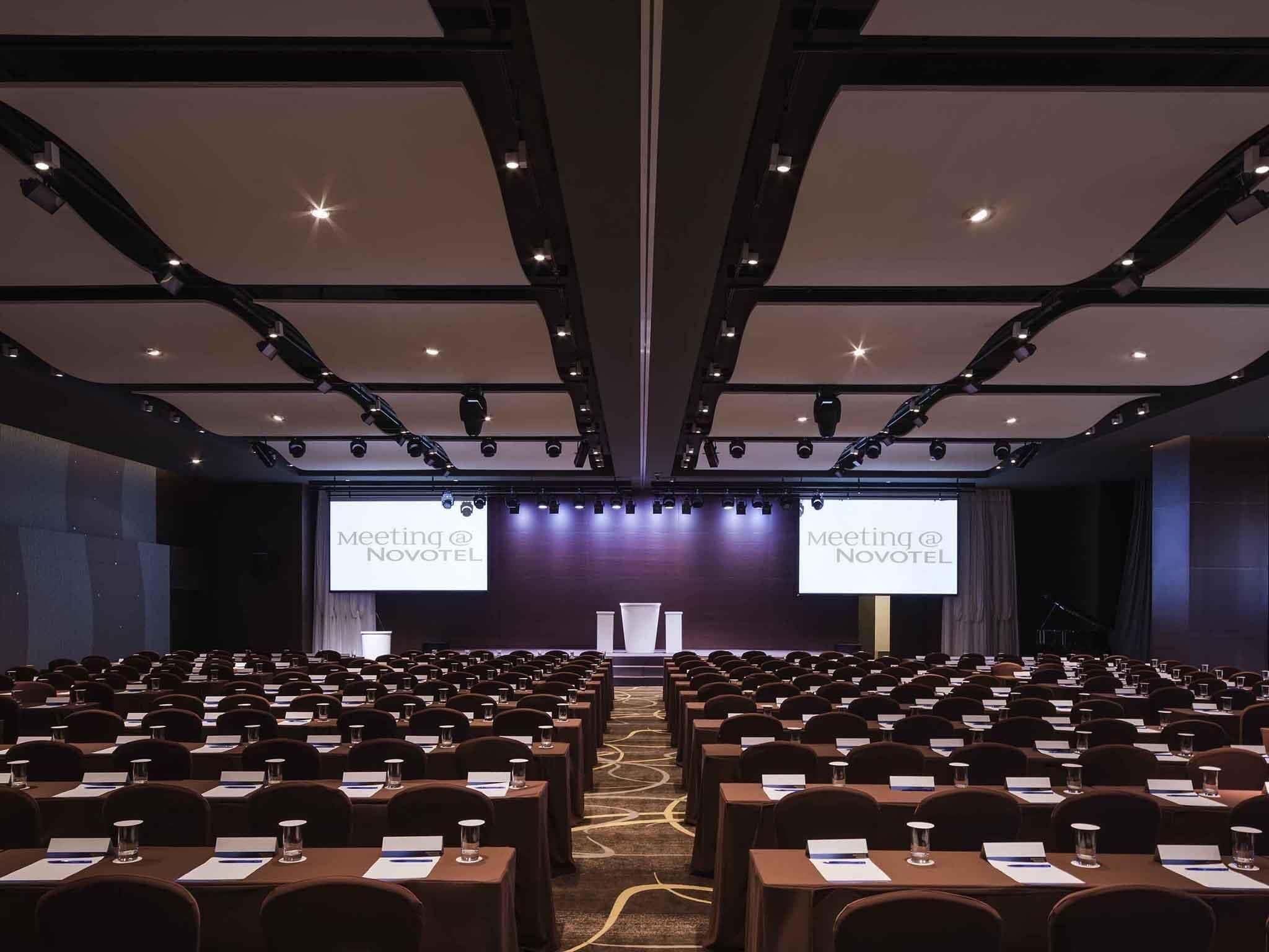 Un salón de conferencias con filas de mesas y sillas, un escenario con un atril y dos pantallas grandes mostrando 'Meeting @ Novotel'.