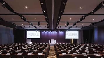 Un salón de conferencias con filas de mesas y sillas, un escenario con un atril y dos pantallas grandes mostrando 'Meeting @ Novotel'.