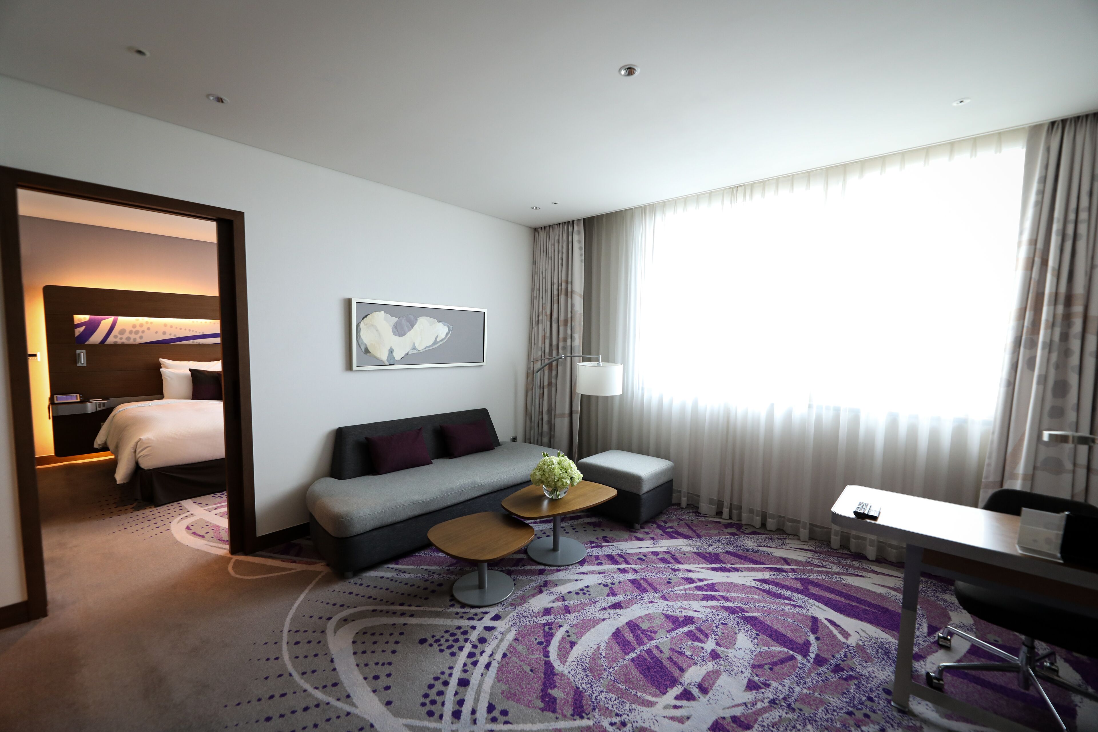 A modern hotel room with a sofa, a small coffee table, a desk, and a large window with curtains.