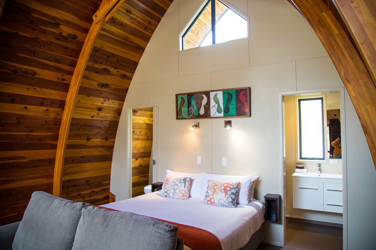 Un dormitorio con techo inclinado, una cama con almohadillas estampadas, una mesita de noche y un baño con ventana y lavamanos.