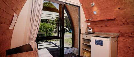 Una habitación compacta con detalles de madera, una puerta corrediza de vidrio que da a un espacio exterior, una pequeña cocina con refrigerador y un televisor de pantalla plana montado en la pared.