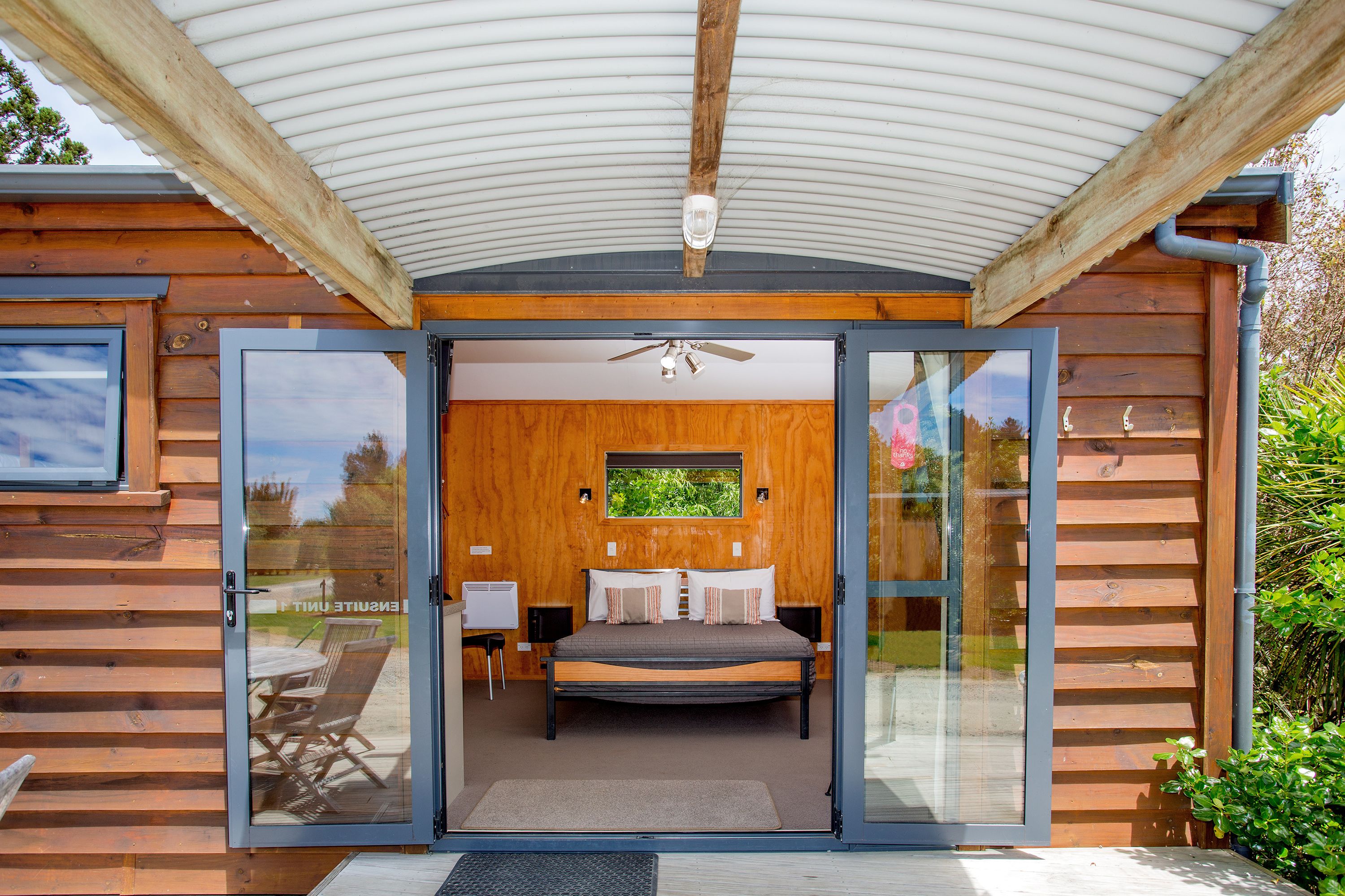 A wooden cabin with a glass door leading to a bedroom with a bed, a ceiling fan, and a window.