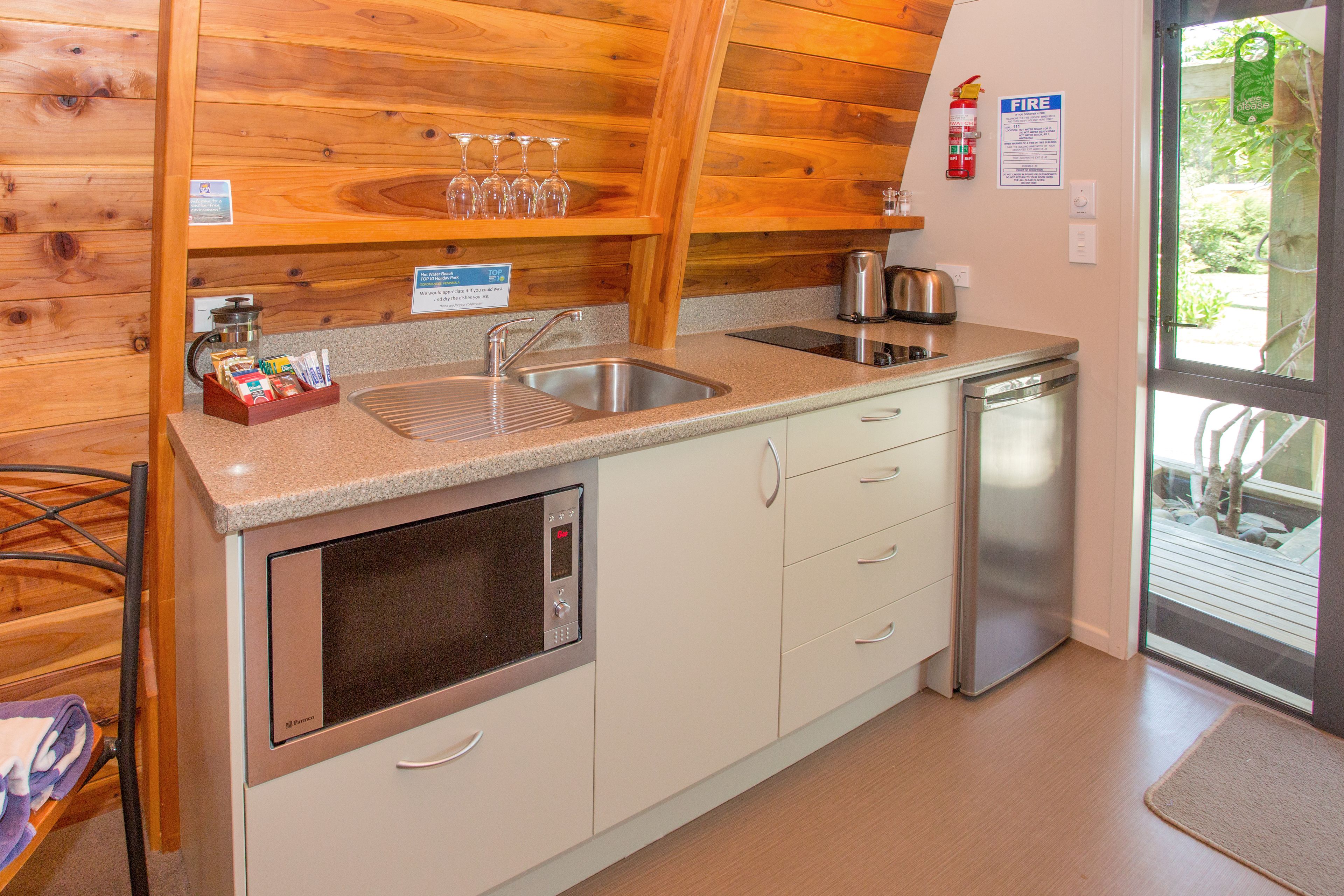 Een compacte keuken met een spoelbak, fornuis en koffiezetapparaat. Er staan wijnglazen op een plank boven de spoelbak.