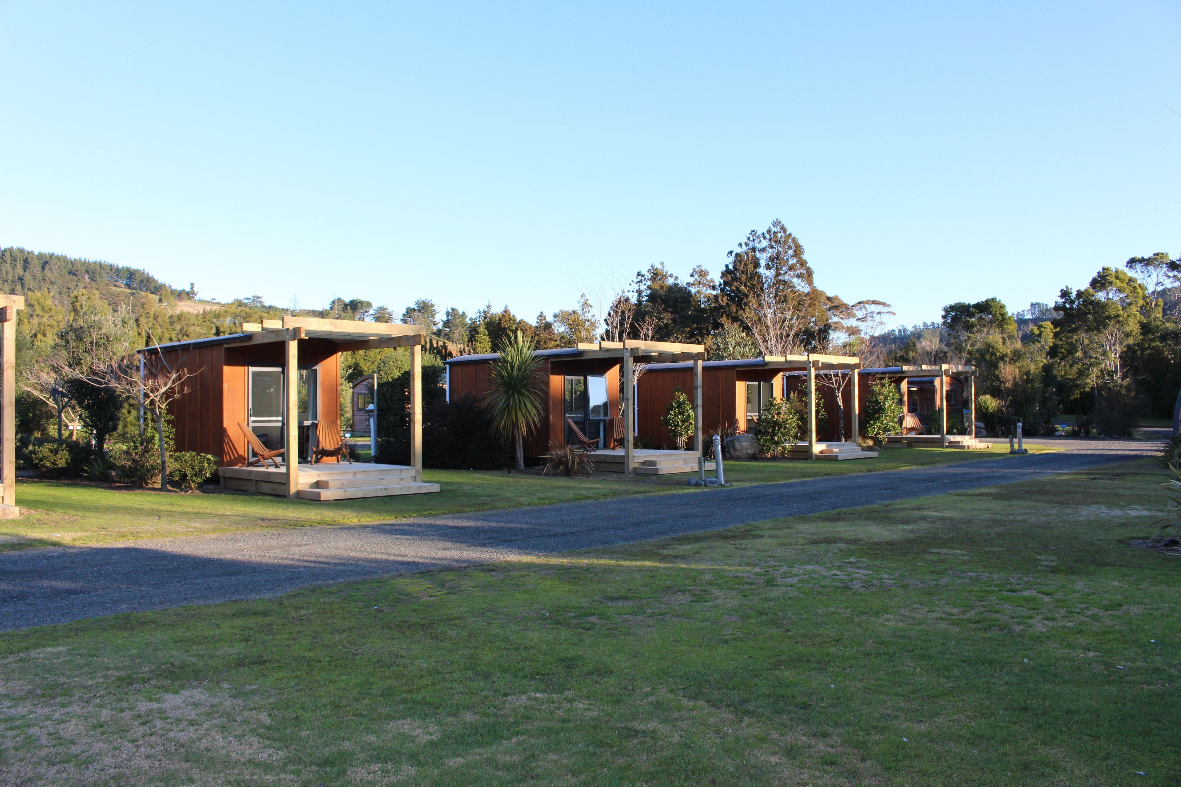 A row of modern, single-story cabins with wooden exteriors and large windows, set in a grassy area with a clear sky and trees in the background.
