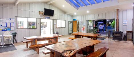 A spacious indoor area with multiple wooden picnic tables and benches, a television mounted on the wall, and a natural light source from skylights.
