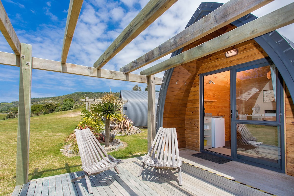 Una terraza de madera con dos sillones de descanso, una pérgola de madera y vista a una casa moderna con puerta de vidrio.