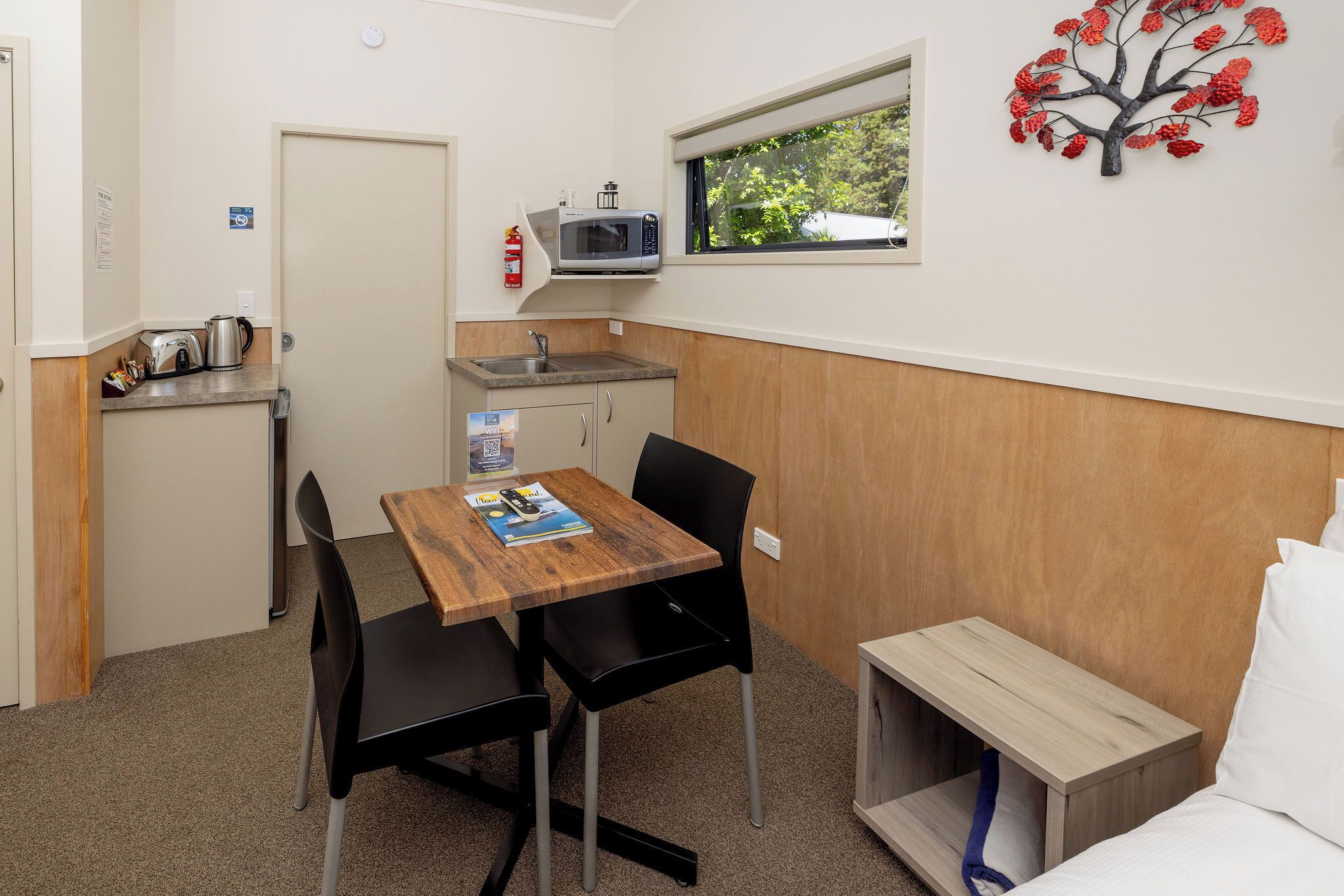 A compact room with a kitchenette, a dining table with chairs, a bed with a wooden headboard, and a window with a view of greenery.