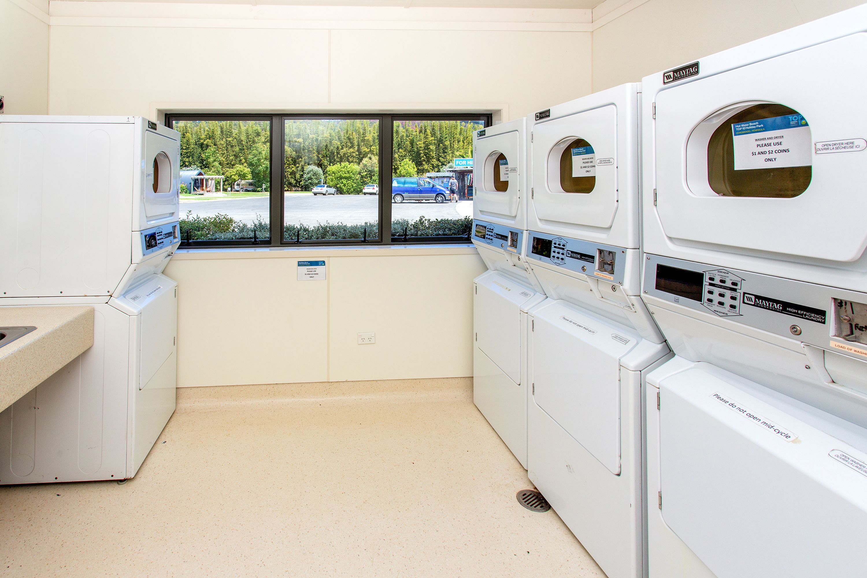 A laundry room with multiple washing machines and dryers, a window with a view of a parking lot and trees, and a sign instructing to use coins for payment.