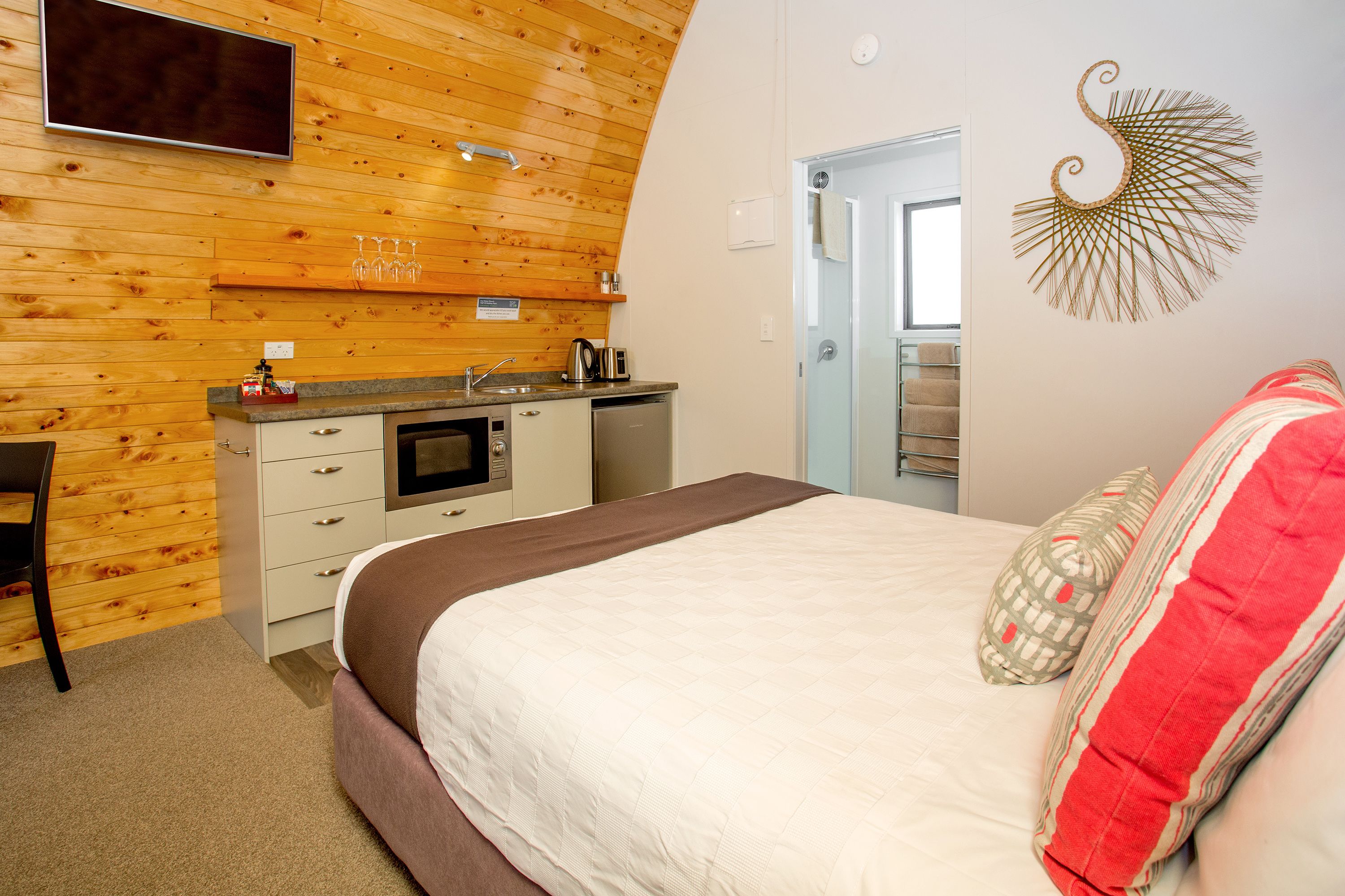 A bedroom with a bed, a desk, a kitchenette, and a door leading to a bathroom.