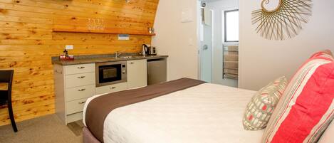 A bedroom with a bed, a desk, a kitchenette, and a door leading to a bathroom.