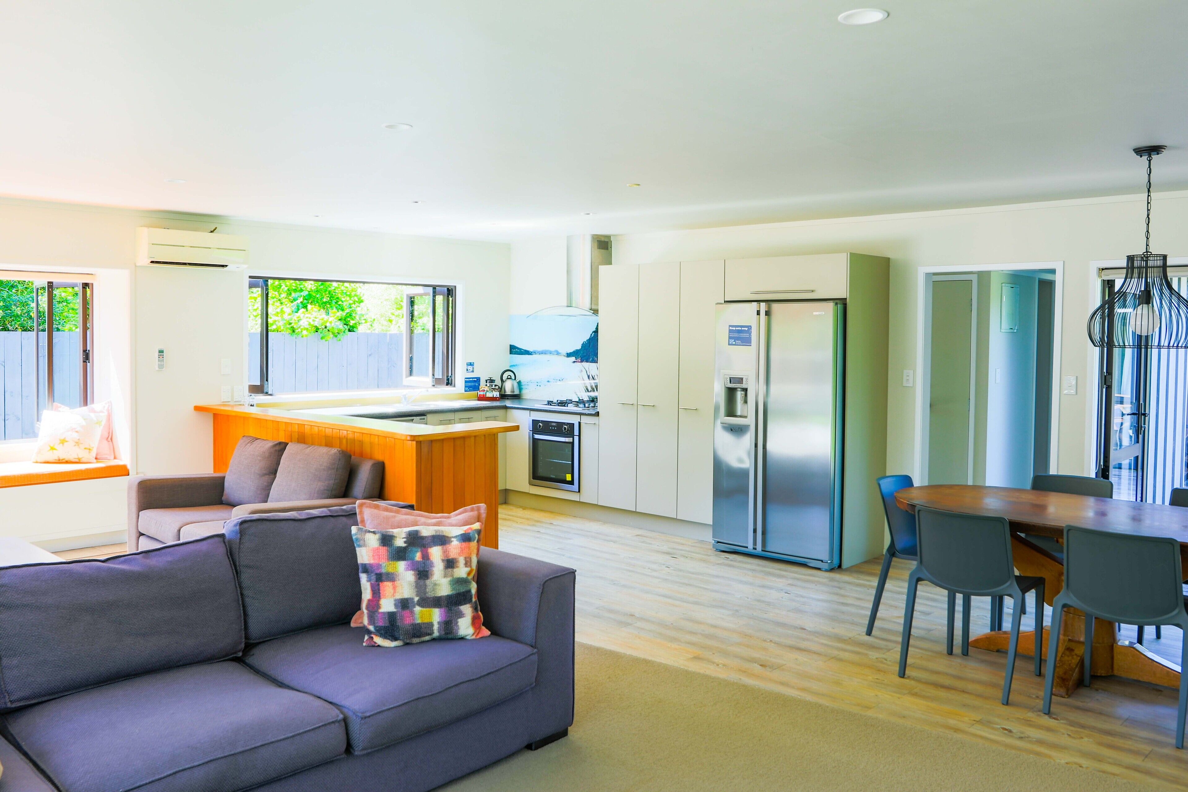 A modern kitchen with a central island, a dining area with a wooden table and chairs, and a living room with a sofa and a colorful cushion.