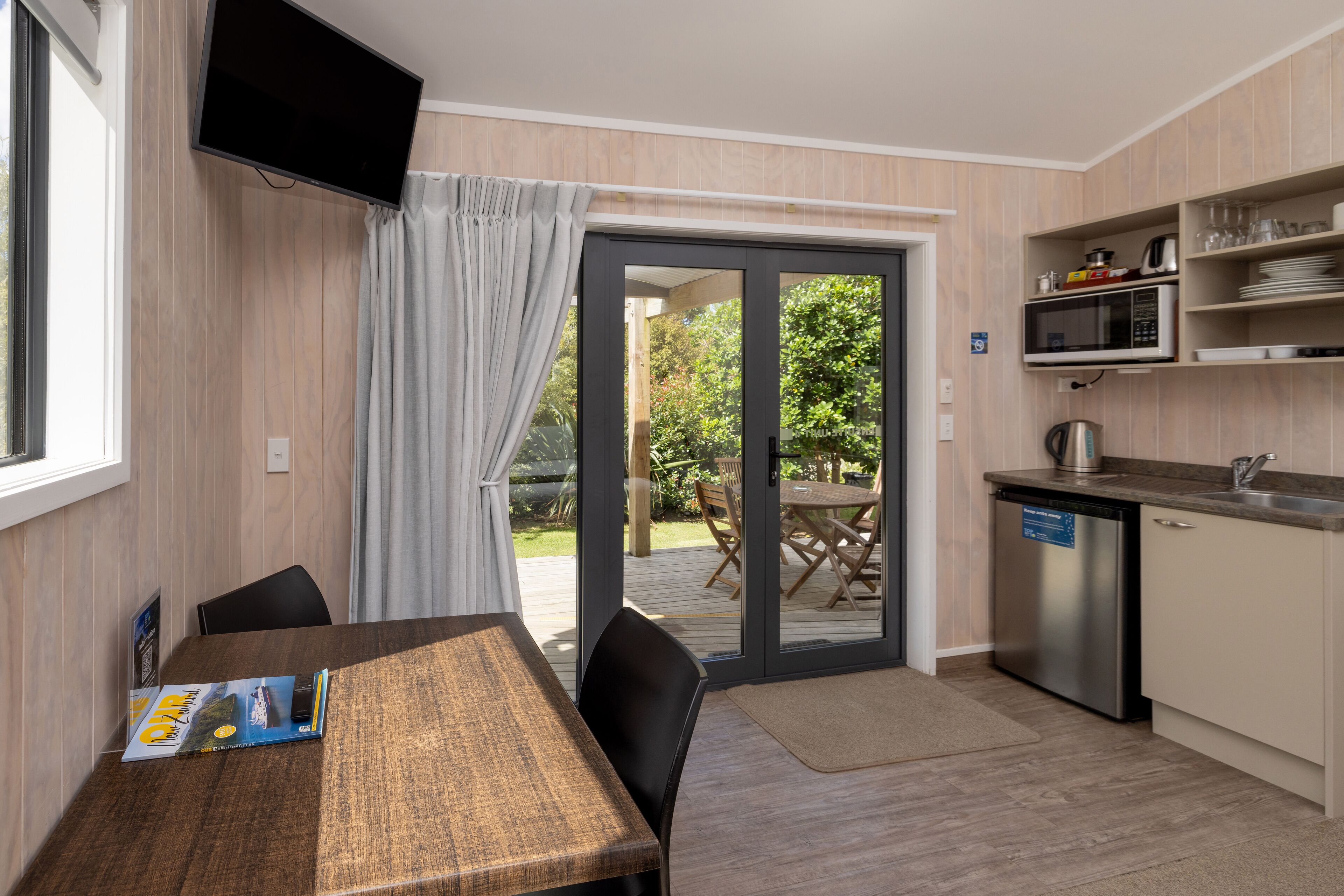 A compact kitchen with a dining area, a TV mounted on the wall, and a sliding glass door leading to an outdoor patio.