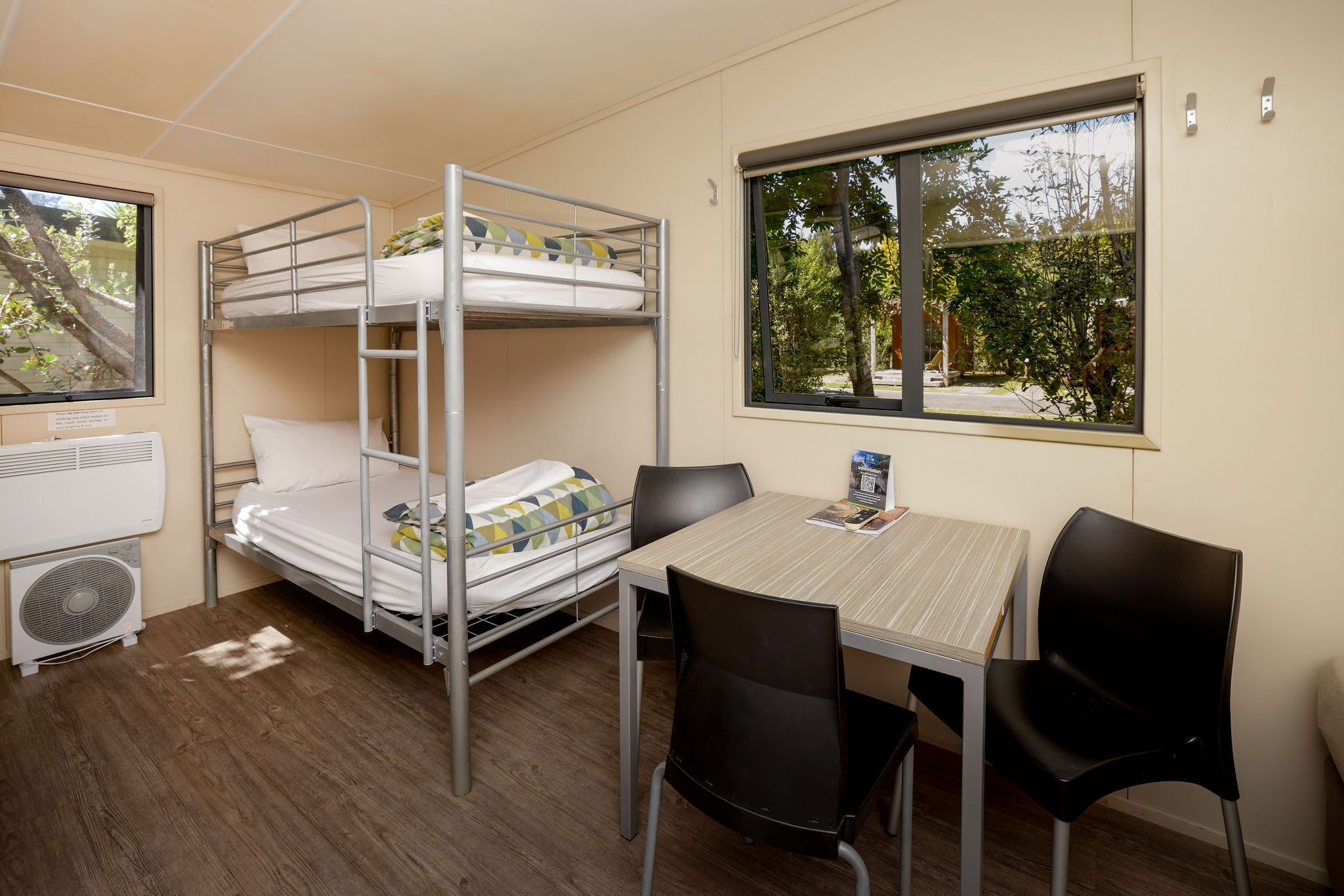 Habitación con literas, una mesa, sillas, un ventanal con vistas a árboles y un ventilador en el suelo.