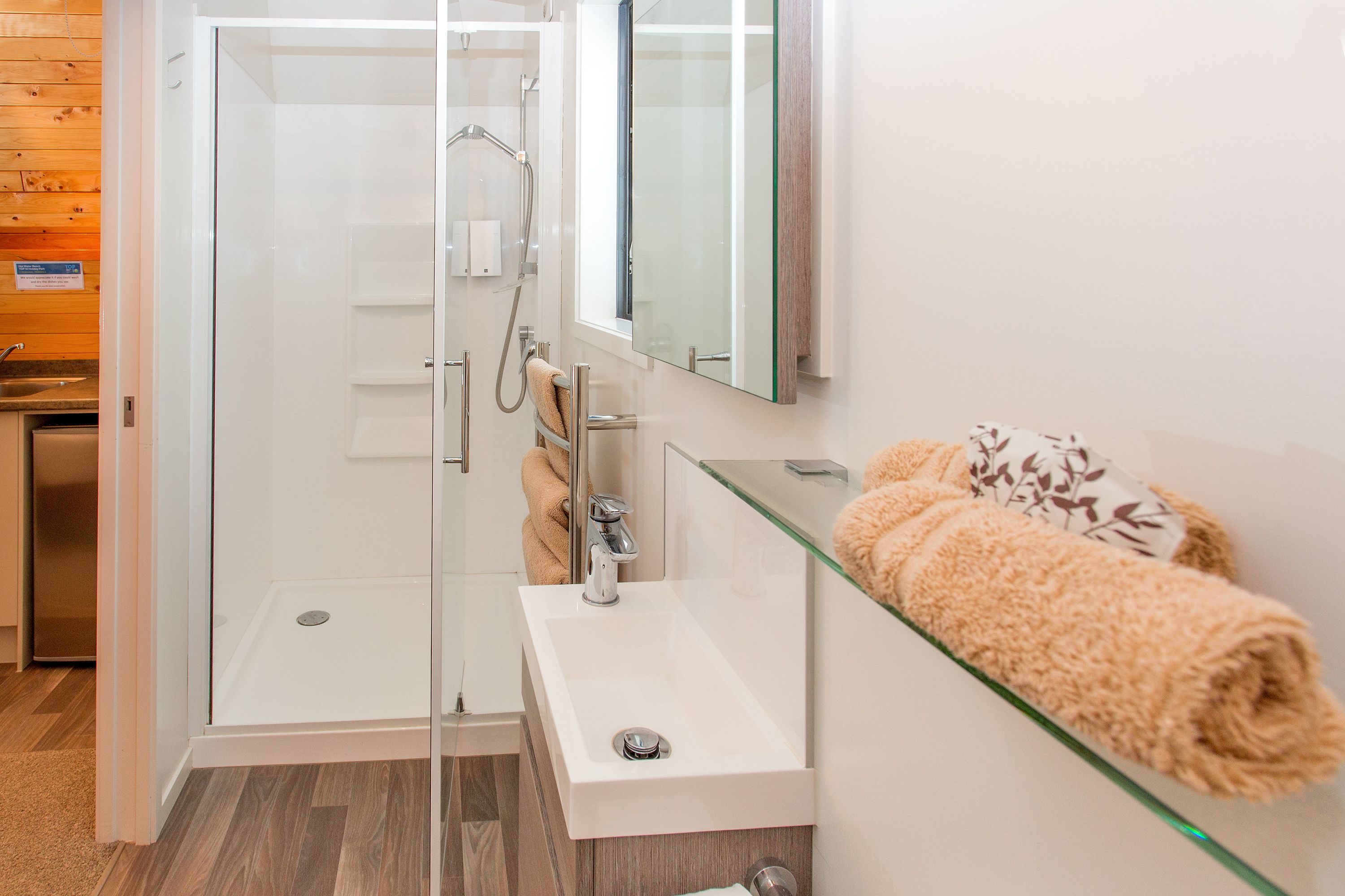 A modern bathroom with a glass-enclosed shower, a white sink, and a glass shelf holding towels.