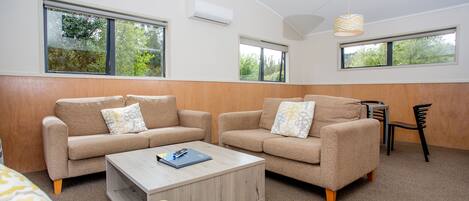 A modern living room with two beige sofas, a wooden coffee table, and a yellow armchair. There are two windows providing natural light and a view of greenery outside.