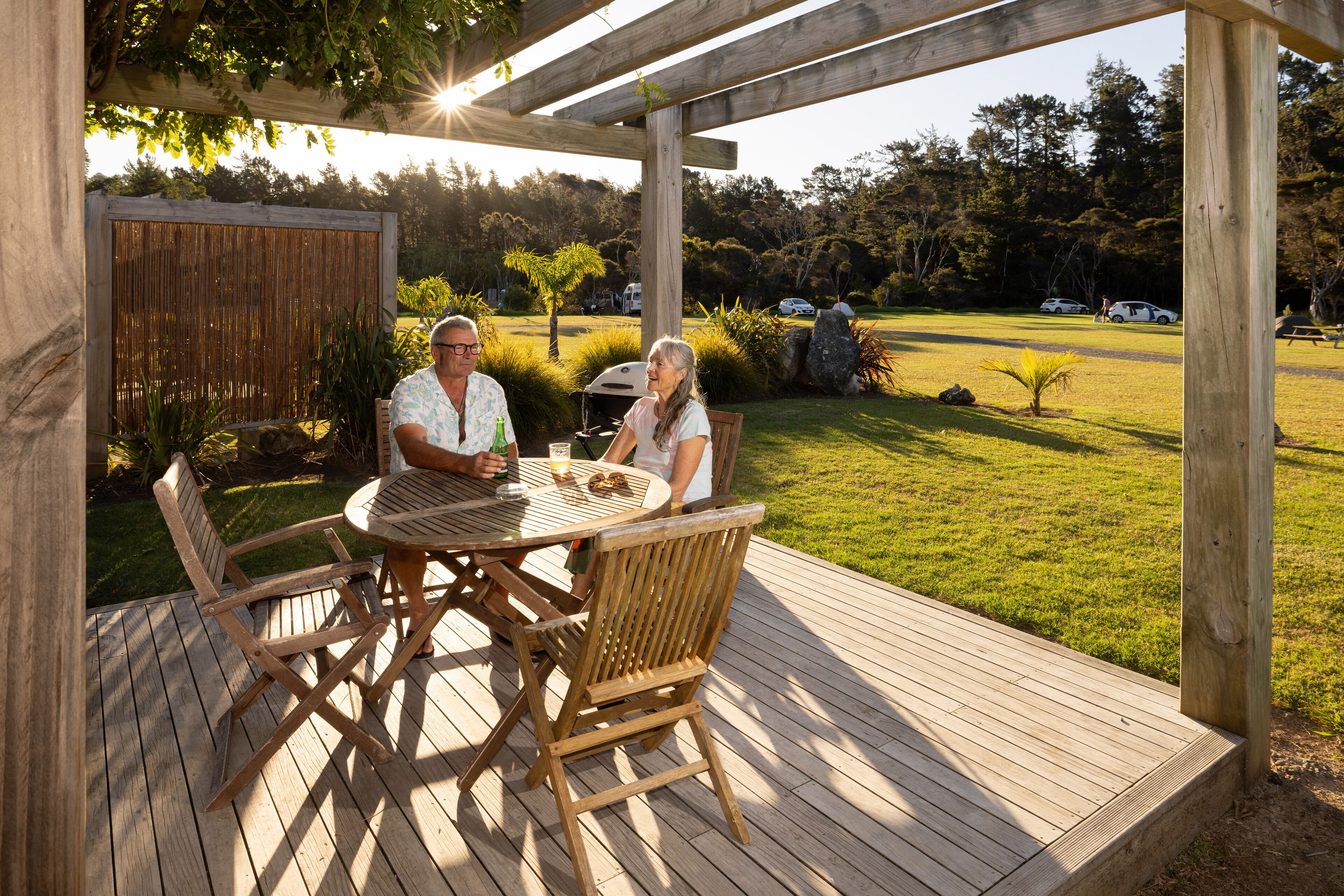 Una terraza de madera con una mesa redonda y sillas, rodeada por una pérgola de madera.