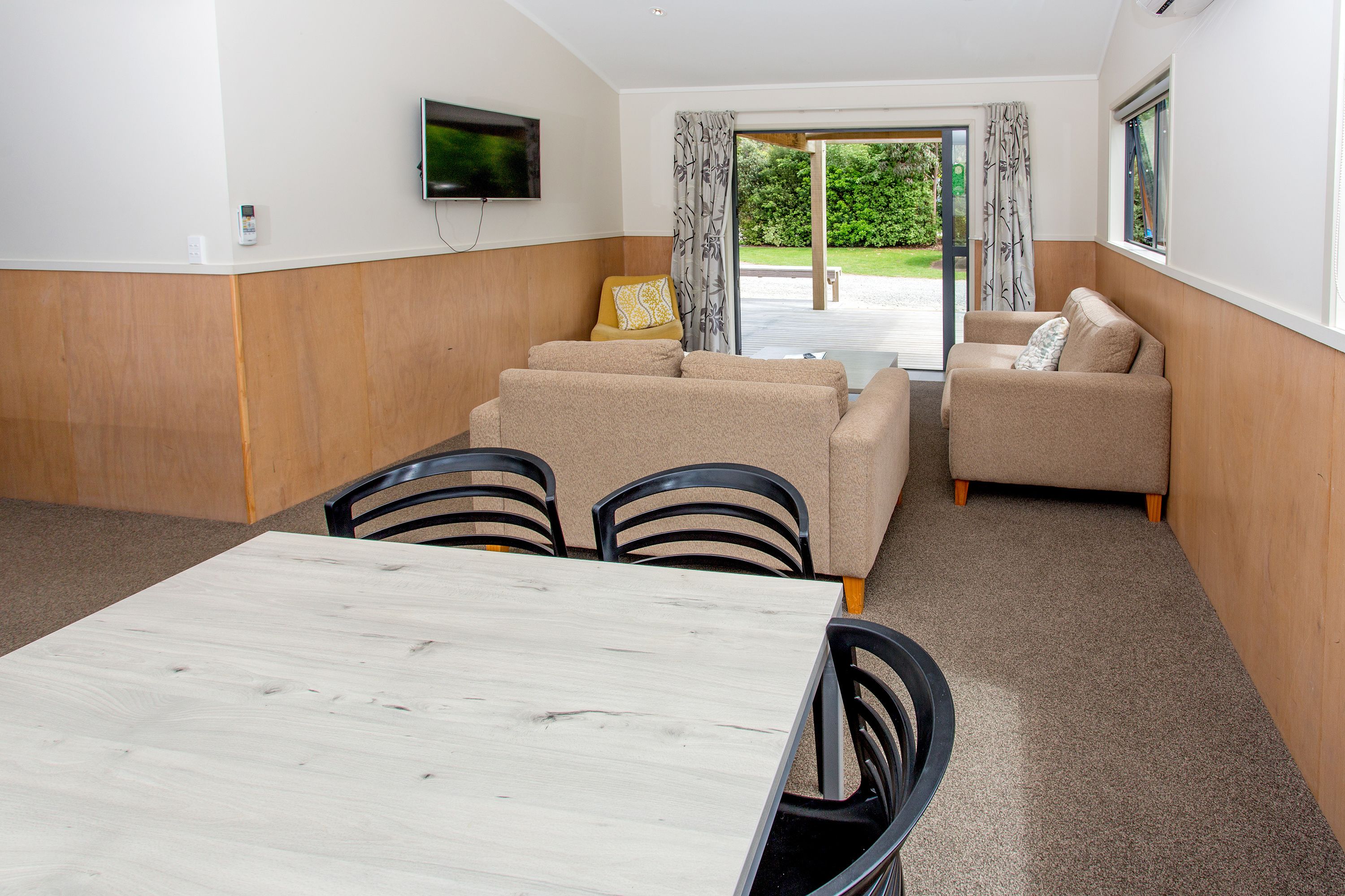 A modern living room with a wooden dining table, beige sofas, and a flat-screen TV mounted on the wall.