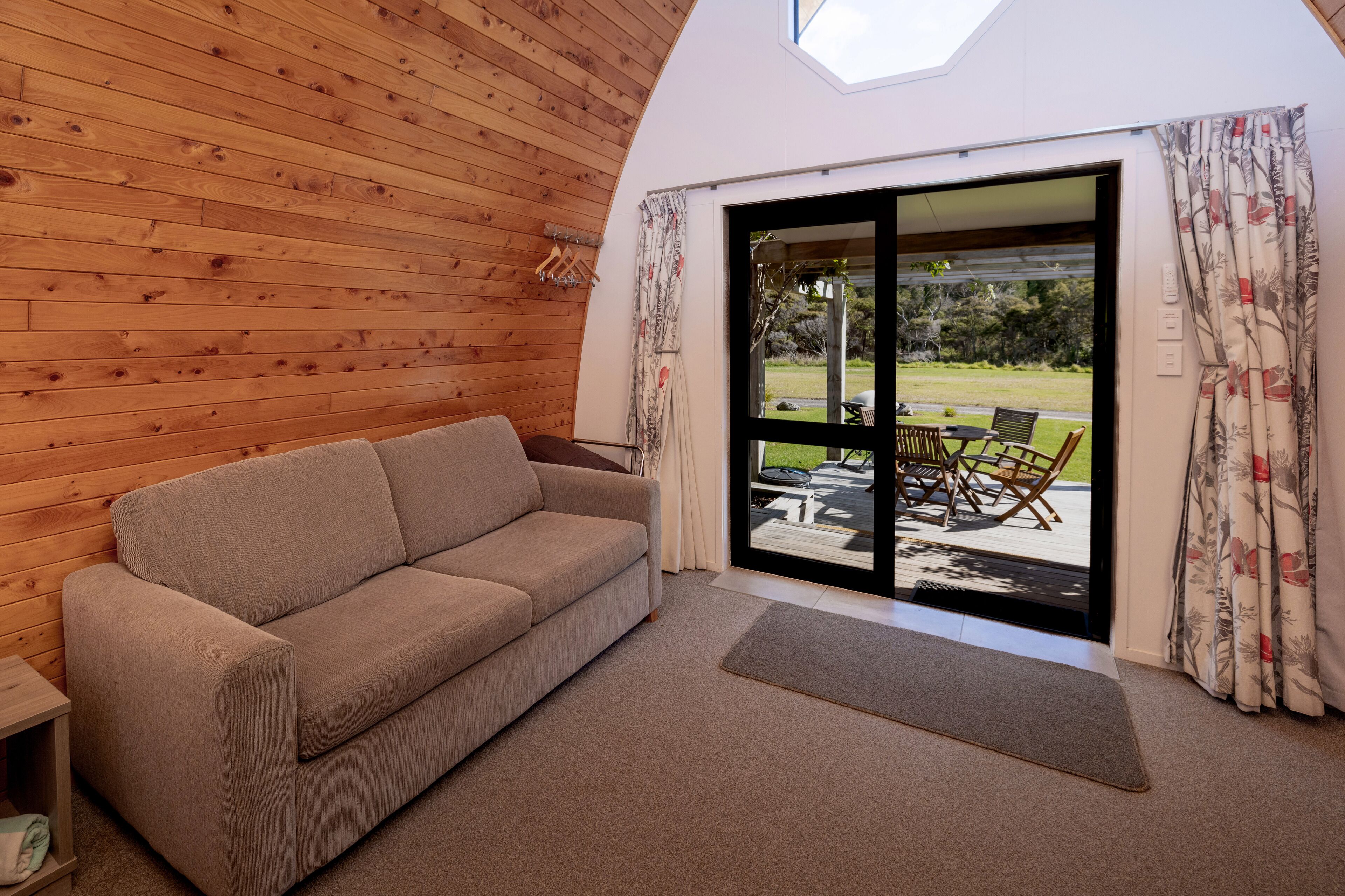 Una habitación acogedora con techo de madera, un sofá color beige, una mesita pequeña y un ventanal corredero que da a un área exterior con mesa y sillas.