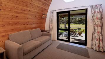 Una habitación acogedora con techo de madera, un sofá color beige, una mesita pequeña y un ventanal corredero que da a un área exterior con mesa y sillas.