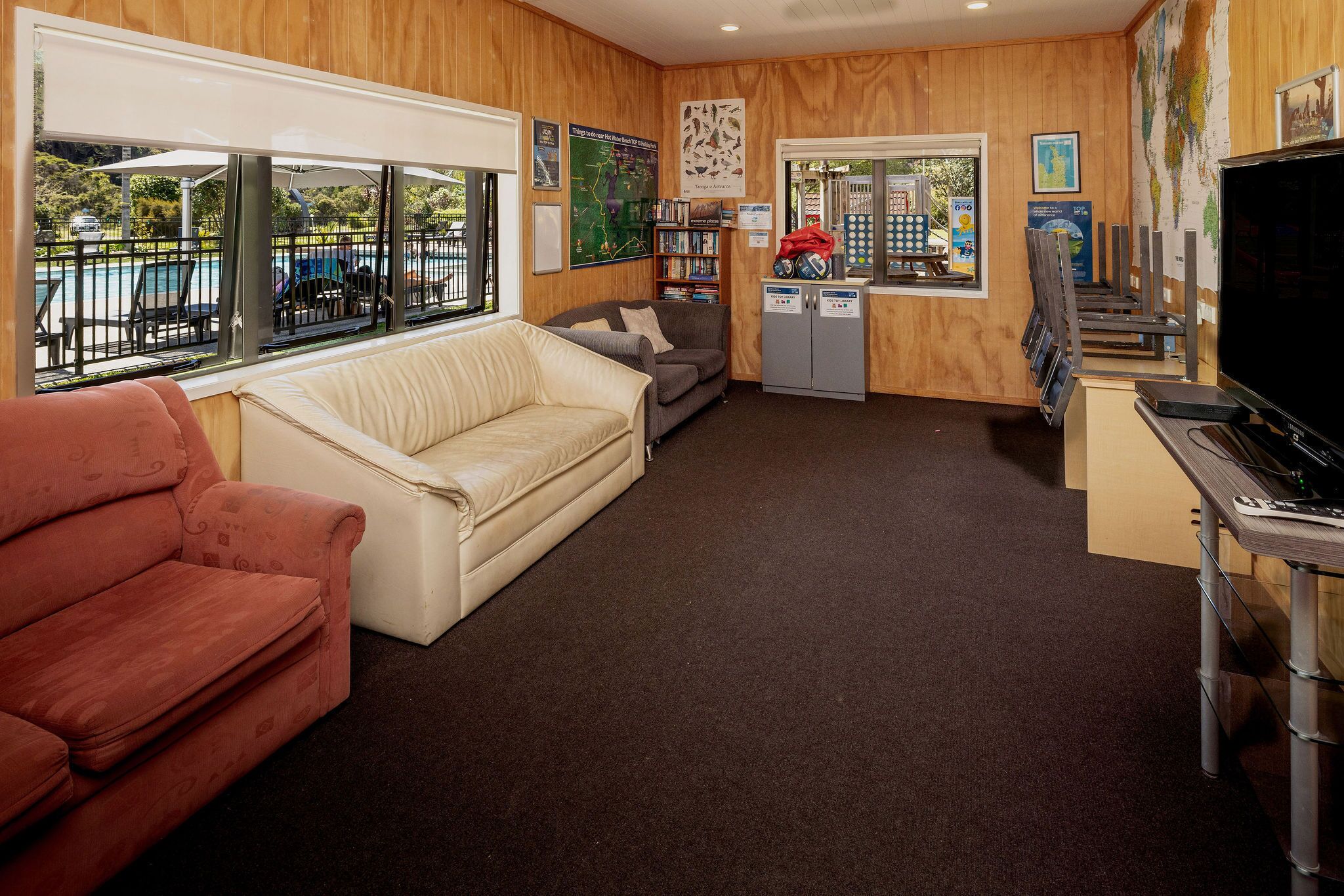 A room with a red sofa, a beige sofa, a grey armchair, a television, a bookshelf, and a window with a view of a pool and greenery.