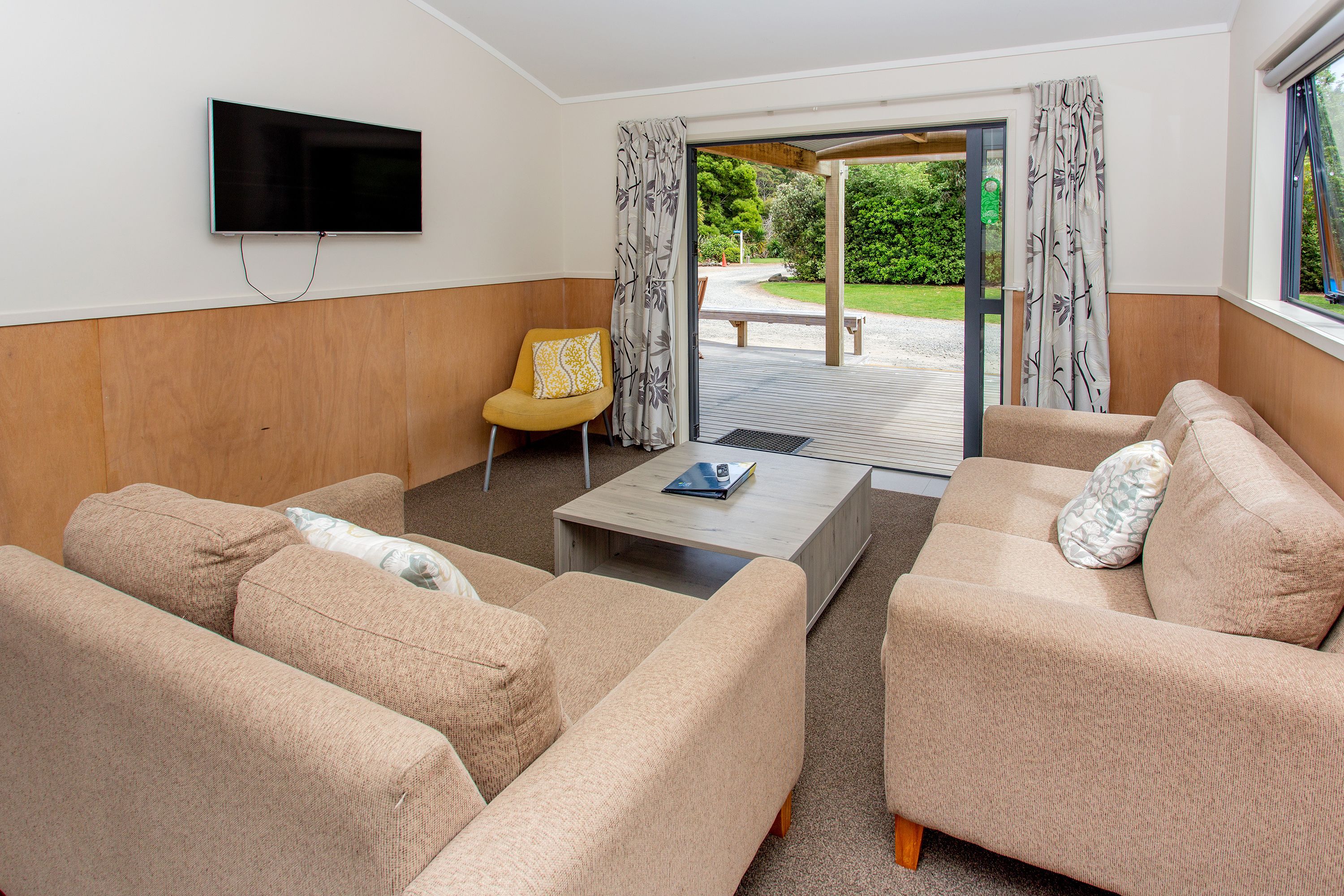 A modern living room with beige sofas, a yellow armchair, a flat-screen TV mounted on the wall, and a wooden coffee table.