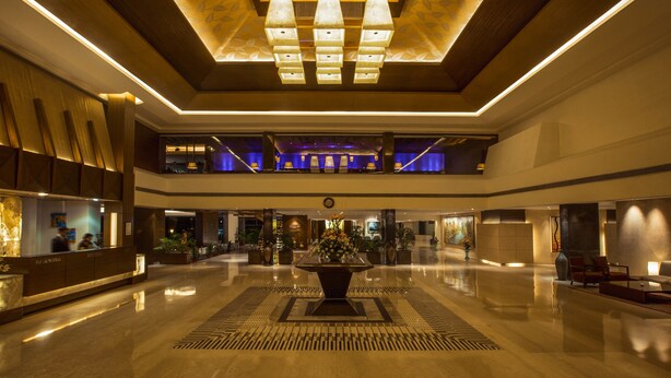 Um hall amplo com uma recepção central, um vaso decorativo grande e um teto com luminária de design geométrico.