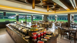 Uma área de buffet com diversos pontos de comida, arranjos de assento e grandes janelas com vista para um campo verde.