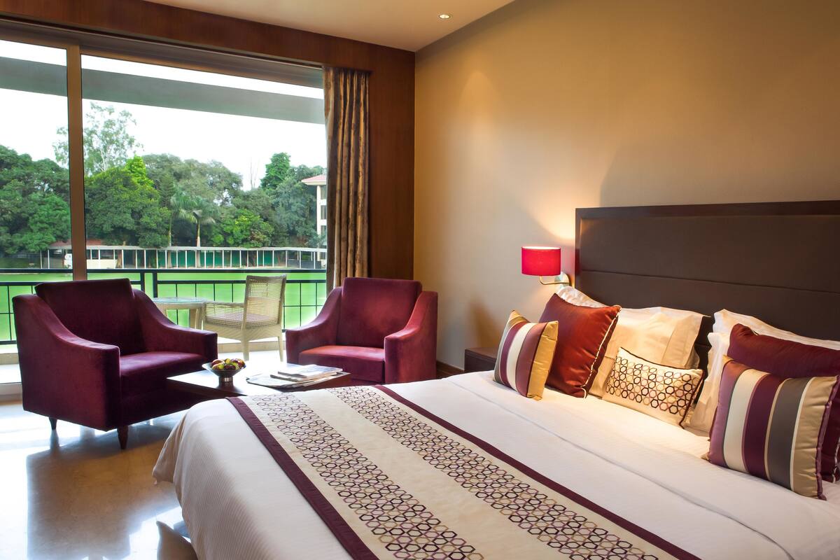 A hotel room with a large bed, two armchairs, a small table with a bowl of fruit, and a view of greenery through large windows.