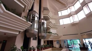 Um átrio espaçoso com um elemento central de água, rodeado por plantas em vasos, tetos altos com padrões geométricos e grandes janelas que permitem a entrada de luz natural.